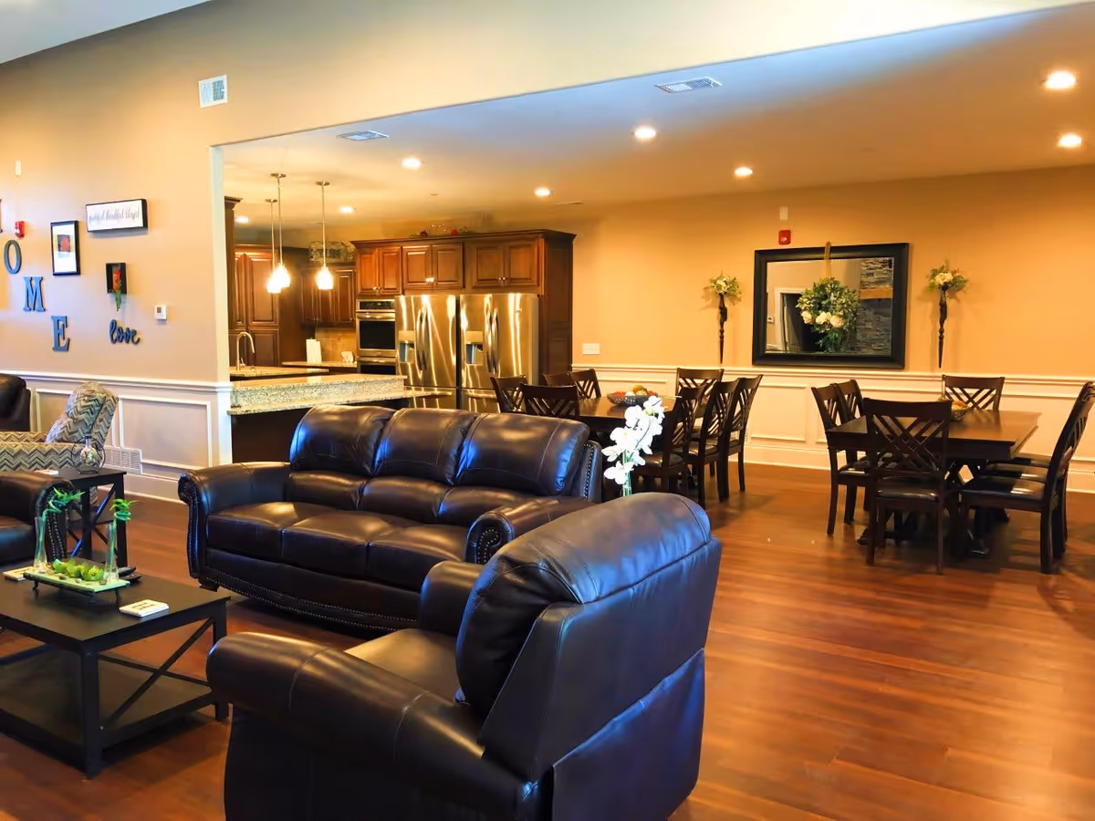 Open senior living common area with dark leather sofas, a coffee table, dining tables and a kitchen in the background.