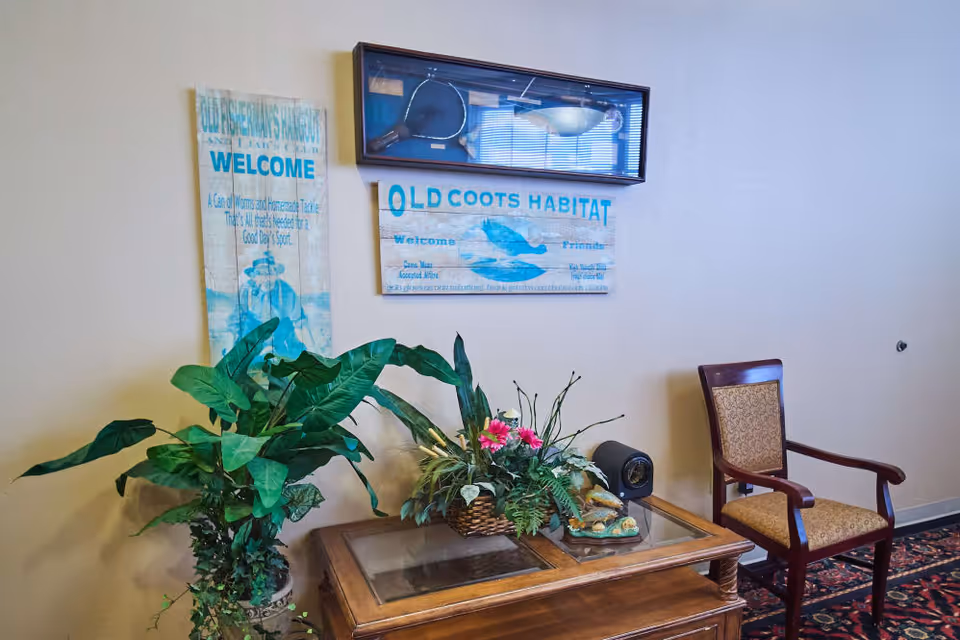 A corner of a room with a wooden table holding a floral arrangement and a small decorative item. Next to the table is a wooden chair with patterned upholstery. On the wall above the table are two rustic-style signs, one reading 'OLD COOTS HABITAT' and the other welcoming to a fishing hangout. A framed display case with fishing gear is mounted above the signs. A large green potted plant is placed to the left of the table.