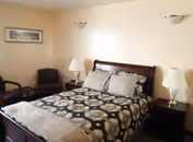 A cozy bedroom with a wooden bed frame, patterned bedding, two bedside tables each with a lamp, two chairs in the corner, and a framed picture on the wall.