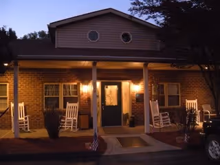 Front exterior view of a single-story brick building at dusk with a covered porch featuring four white rocking chairs, two lit wall-mounted lanterns flanking a central door, small American flags near the entrance, and some trees partially visible on the right side.