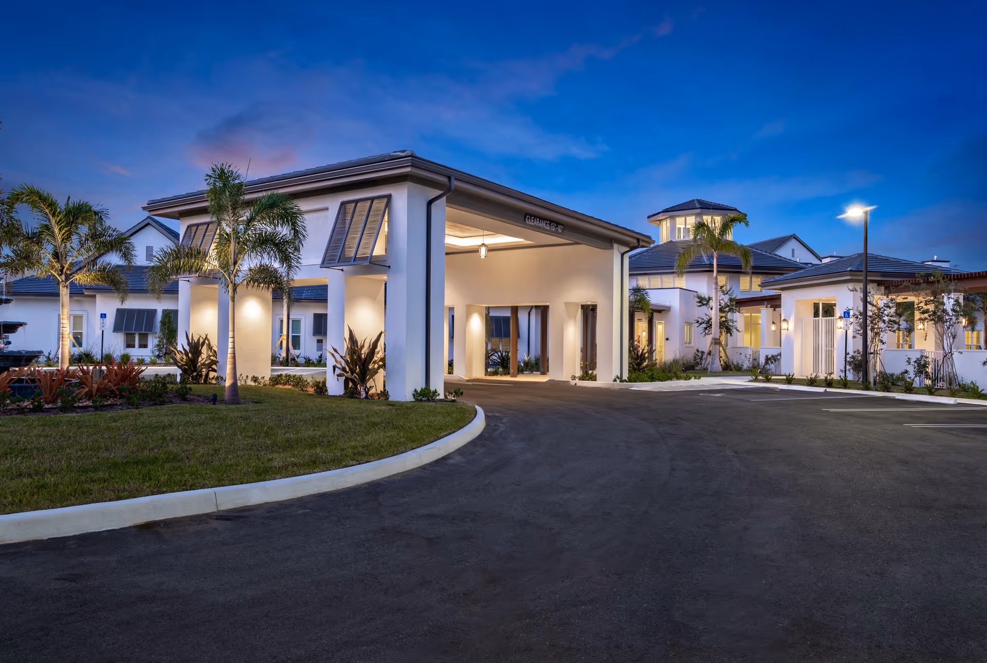 Exterior view of Watercrest St Lucie West facility at dusk, showing a modern building entrance with a covered drop-off area, palm trees, landscaped greenery, and a clear sky with soft lighting.