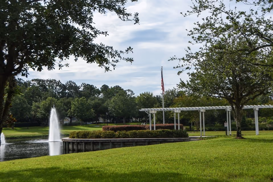 A peaceful outdoor scene at Westminster St. Augustine featuring a well-maintained grassy area, a pond with two water fountains, an American flag on a flagpole, and a white pergola structure surrounded by trees and shrubs under a partly cloudy sky.