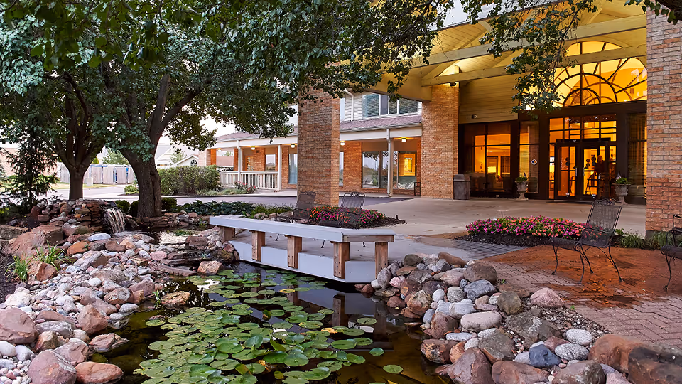Covered brick entrance to a senior living facility beside a landscaped pond with lily pads and a small wooden footbridge.
