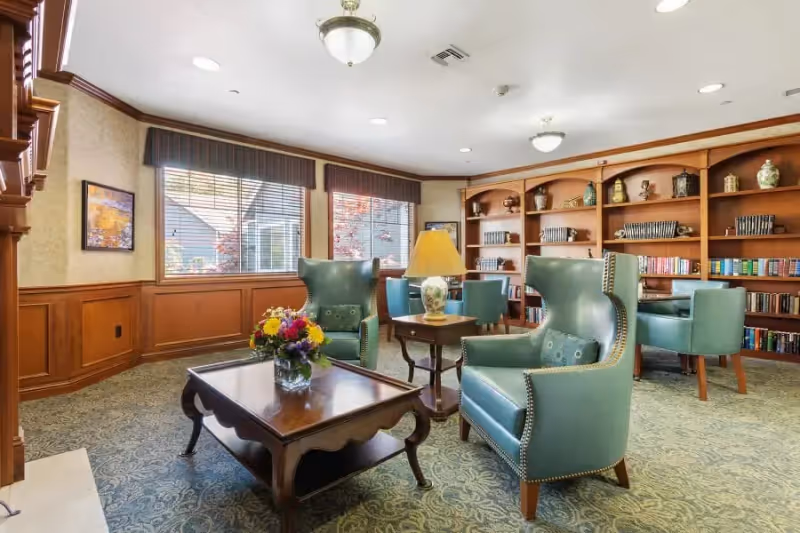 A cozy sitting area in a senior living facility with two teal wingback chairs and a wooden coffee table with a flower arrangement. Behind the chairs is a wooden bookshelf filled with books and decorative items. The room has large windows with blinds and valances, wood paneling on the walls, and a patterned carpet. A table lamp sits on a small side table between the chairs.