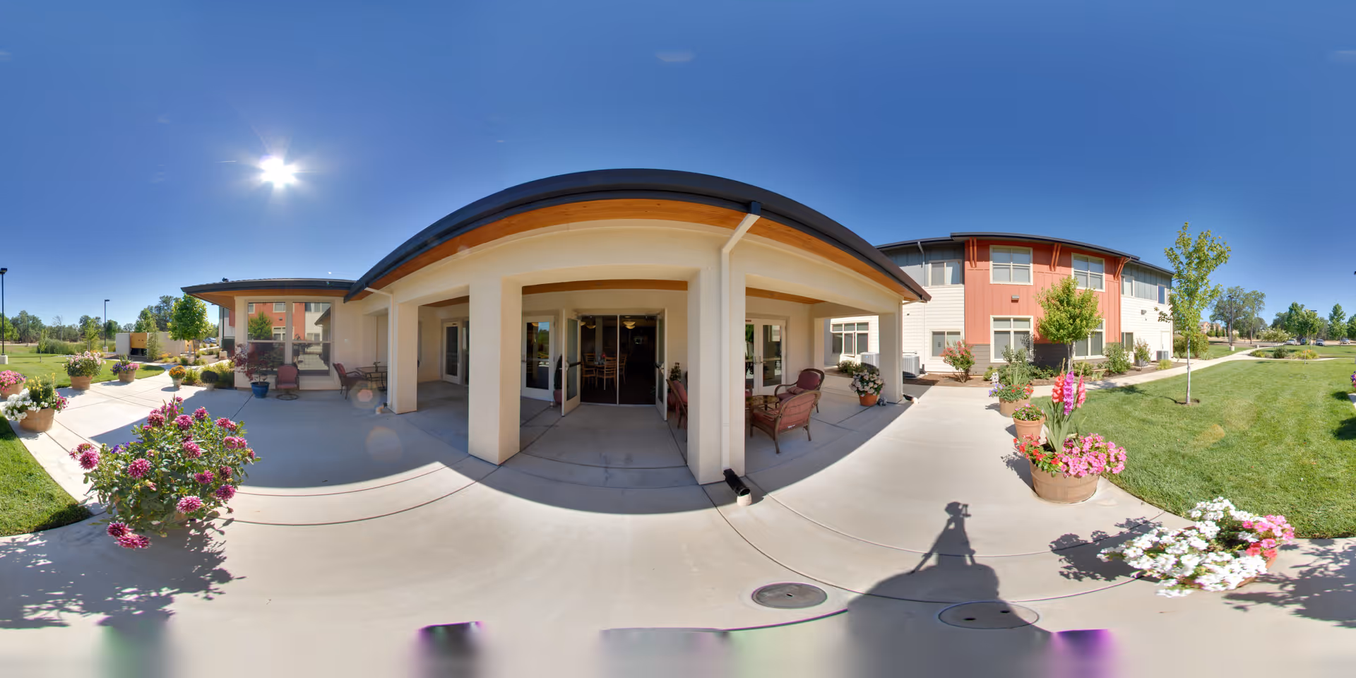 Outdoor patio area of The Vistas Assisted Living and Memory Care facility on a sunny day, featuring a covered seating area with chairs and potted plants, surrounded by well-maintained green lawns and flower beds under a clear blue sky.