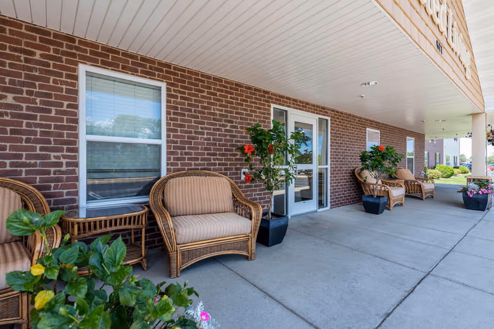 Covered outdoor seating area with wicker chairs and small tables along a brick wall. There are potted plants with red flowers placed between the chairs. A glass door and windows are visible on the brick wall, and the area is paved with concrete.
