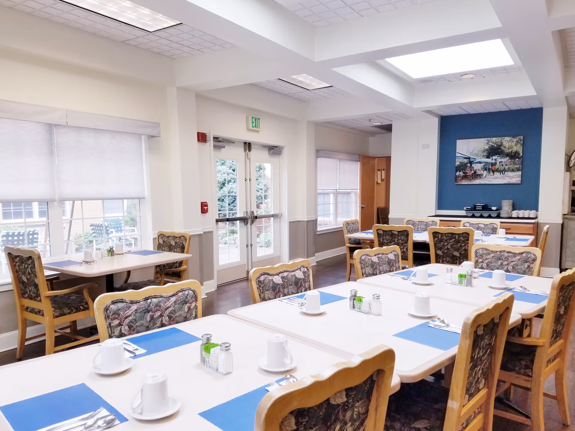 A bright dining room with multiple tables and chairs arranged neatly. Each table is set with white cups, saucers, silverware, salt and pepper shakers, and blue placemats. The room has large windows with blinds, a double glass door leading outside, and a painting on a blue accent wall. The chairs have wooden frames and floral-patterned cushions.