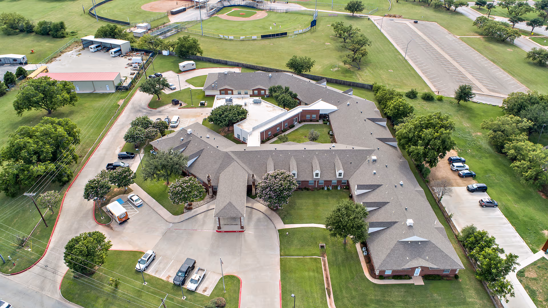 Aerial view of Morada Abilene senior living facility showing a large, single-story building with a gray roof surrounded by green lawns, trees, parking areas, and nearby baseball fields.