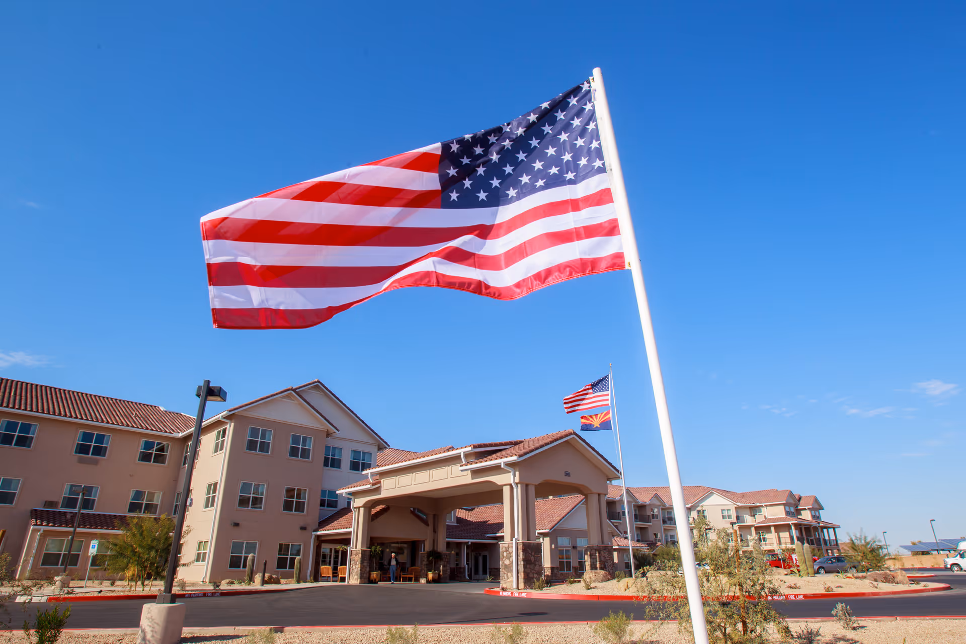 Exterior view of Estrella Estates Gracious Retirement Living facility with a large American flag in the foreground and the building with multiple windows and a covered entrance in the background under a clear blue sky.