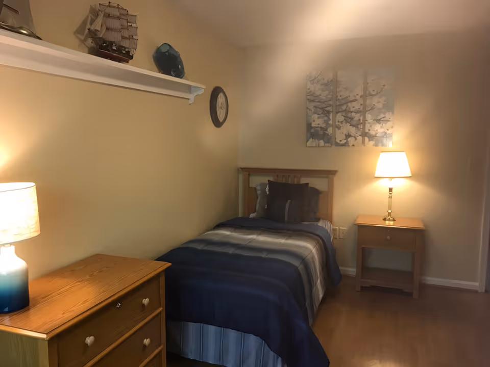 A small bedroom with a single bed covered in a blue and gray striped comforter and multiple pillows. There is a wooden nightstand with a lamp on the right side of the bed, and a wooden dresser with a lamp on the left side. Above the bed is a three-panel wall art featuring white flowers on branches. A small shelf on the left wall holds a model ship and a decorative item, and a round clock is mounted on the wall.