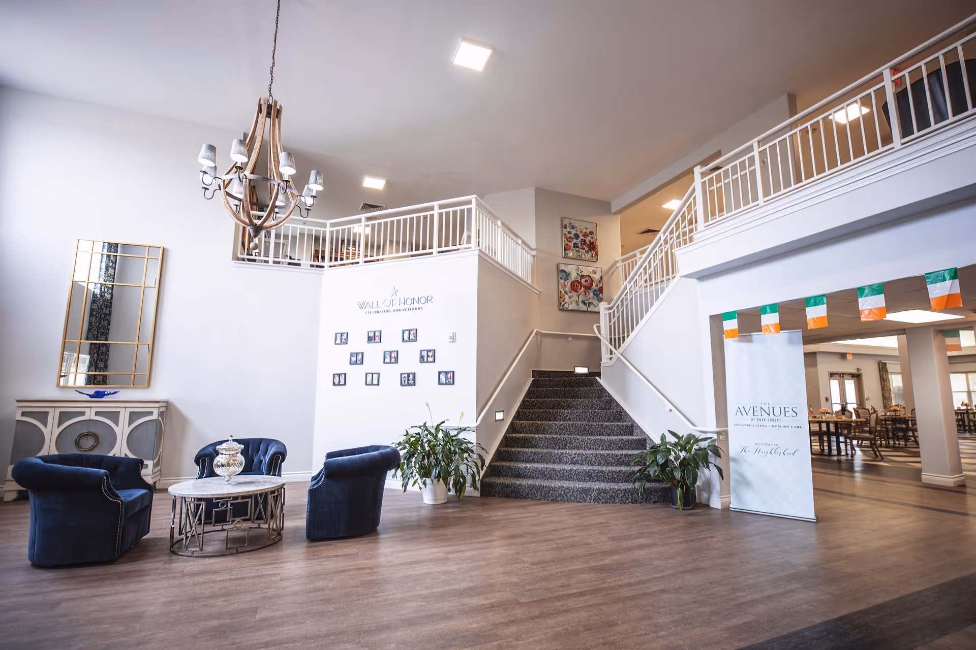 Interior view of a senior living facility lobby with a seating area consisting of three blue armchairs around a round coffee table. A large mirror hangs on the wall above a decorative cabinet. A staircase with white railings leads to an upper floor. On the wall near the stairs is a 'Wall of Honor' display with photos. To the right, a banner reads 'The Avenues at The Grove' with a dining area visible in the background. The space is well-lit with modern ceiling lights and decorated with plants and colorful artwork.