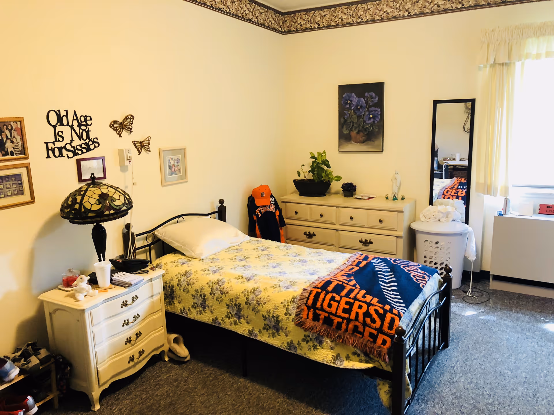 A bright single bedroom with a metal-framed twin bed covered by floral bedding and a Tigers blanket, a nightstand with a lamp, a dresser with plants and a mirror, wall decorations, and a window.