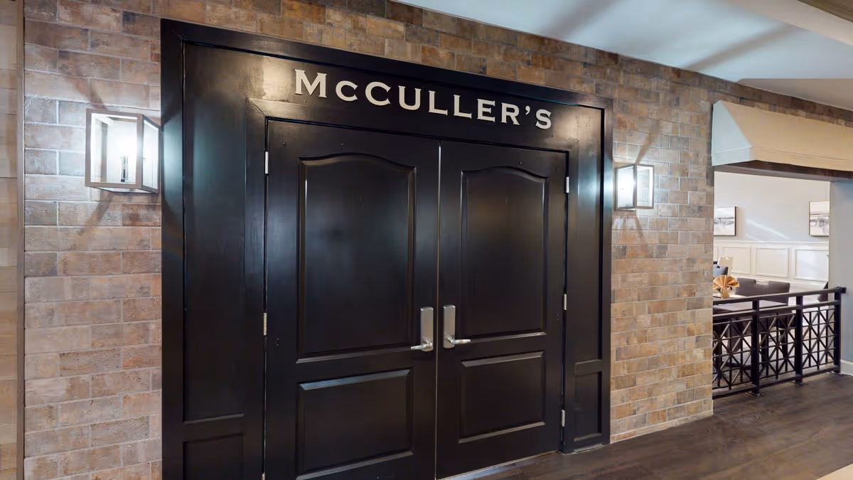Double black doors with silver handles labeled 'McCULLER'S' above them, set in a brick wall with two modern wall sconces on either side. To the right, there is a glimpse of a dining area with tables and chairs.