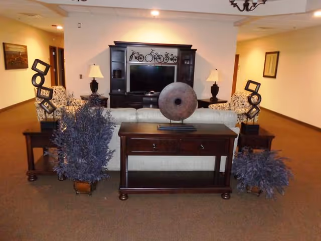 A cozy living room area with a beige sofa facing a dark wooden entertainment center holding a flat-screen TV. Two matching armchairs with patterned upholstery flank the sofa. There are two table lamps on side tables next to the armchairs. Decorative items including sculptures and potted plants are placed on and around a wooden console table behind the sofa. The walls are light-colored with framed artwork, and the carpet is brown.