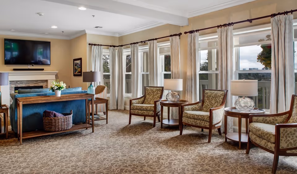 A spacious and well-lit living room with large windows covered by light curtains, several patterned armchairs arranged around wooden side tables with lamps, a blue sofa with a basket underneath, a fireplace with a mounted flat-screen TV above it, and a framed picture on the wall.