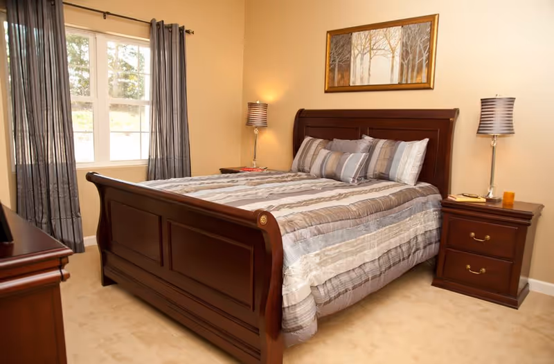A neatly made bed with a wooden frame and striped bedding in a bedroom. There are two matching wooden nightstands on either side of the bed, each with a lamp. A window with gray curtains is on the left wall, and a framed artwork depicting trees hangs above the bed.