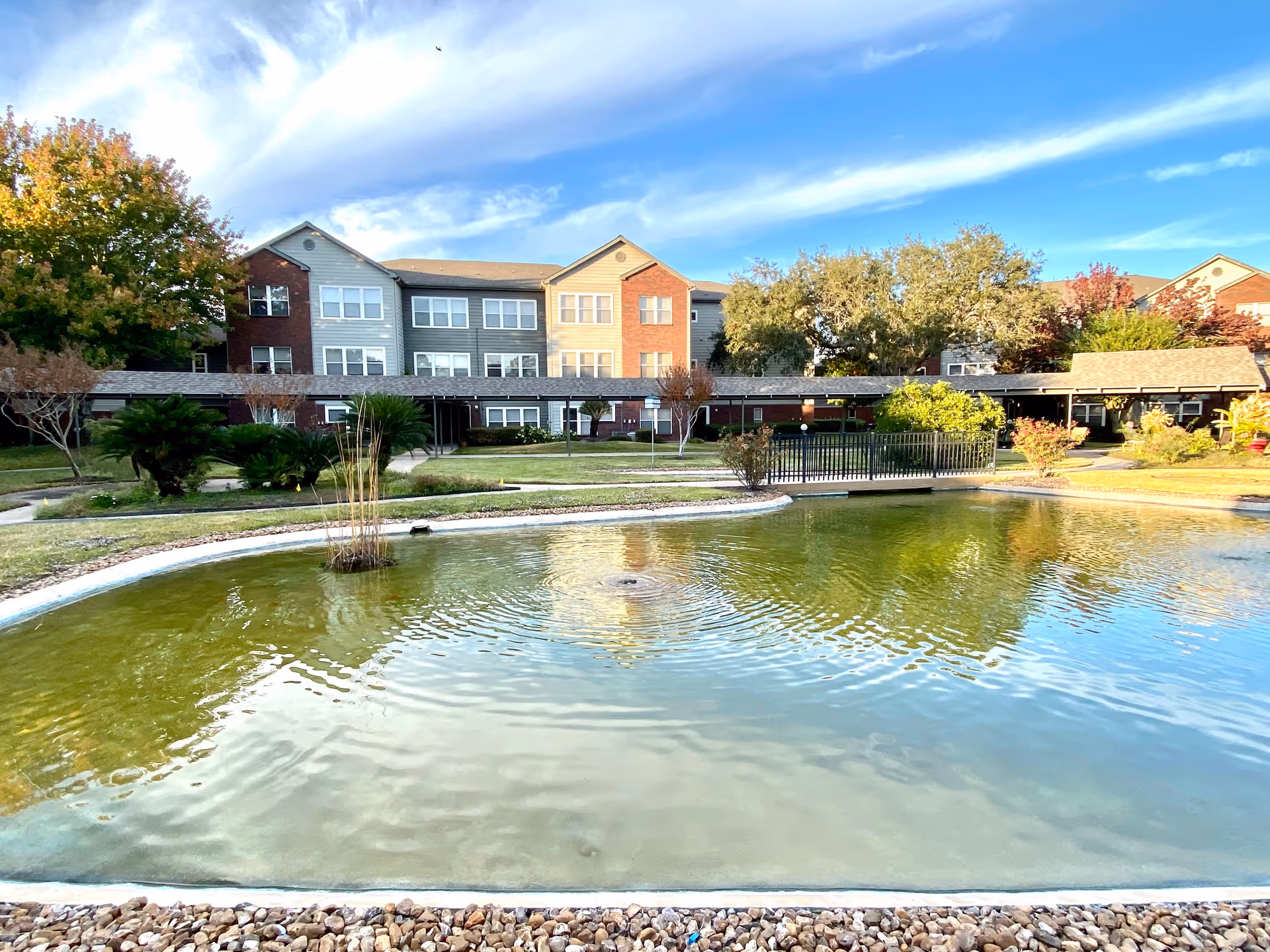 A serene outdoor view of Lone Star Living facility featuring a large pond with a small fountain in the center, surrounded by a stone border. In the background, there is a three-story residential building with a mix of brick and siding exterior, along with well-maintained landscaping including trees, bushes, and a small bridge over the pond.