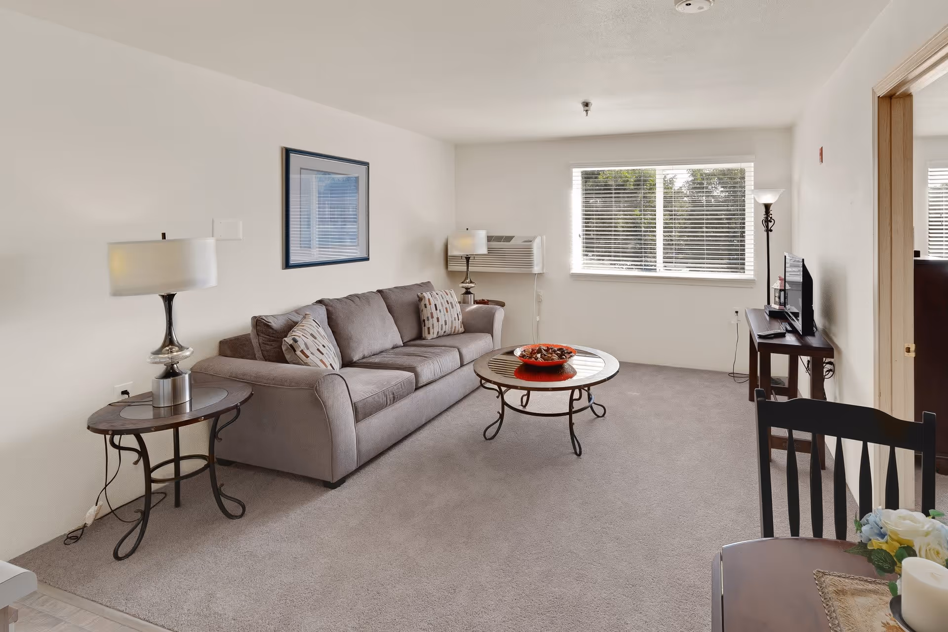 A bright living room with a gray sofa adorned with patterned pillows, a round glass coffee table with a red bowl centerpiece, two side tables each with a lamp, a window with blinds letting in natural light, a TV on a stand, and a dining table with chairs partially visible in the foreground.
