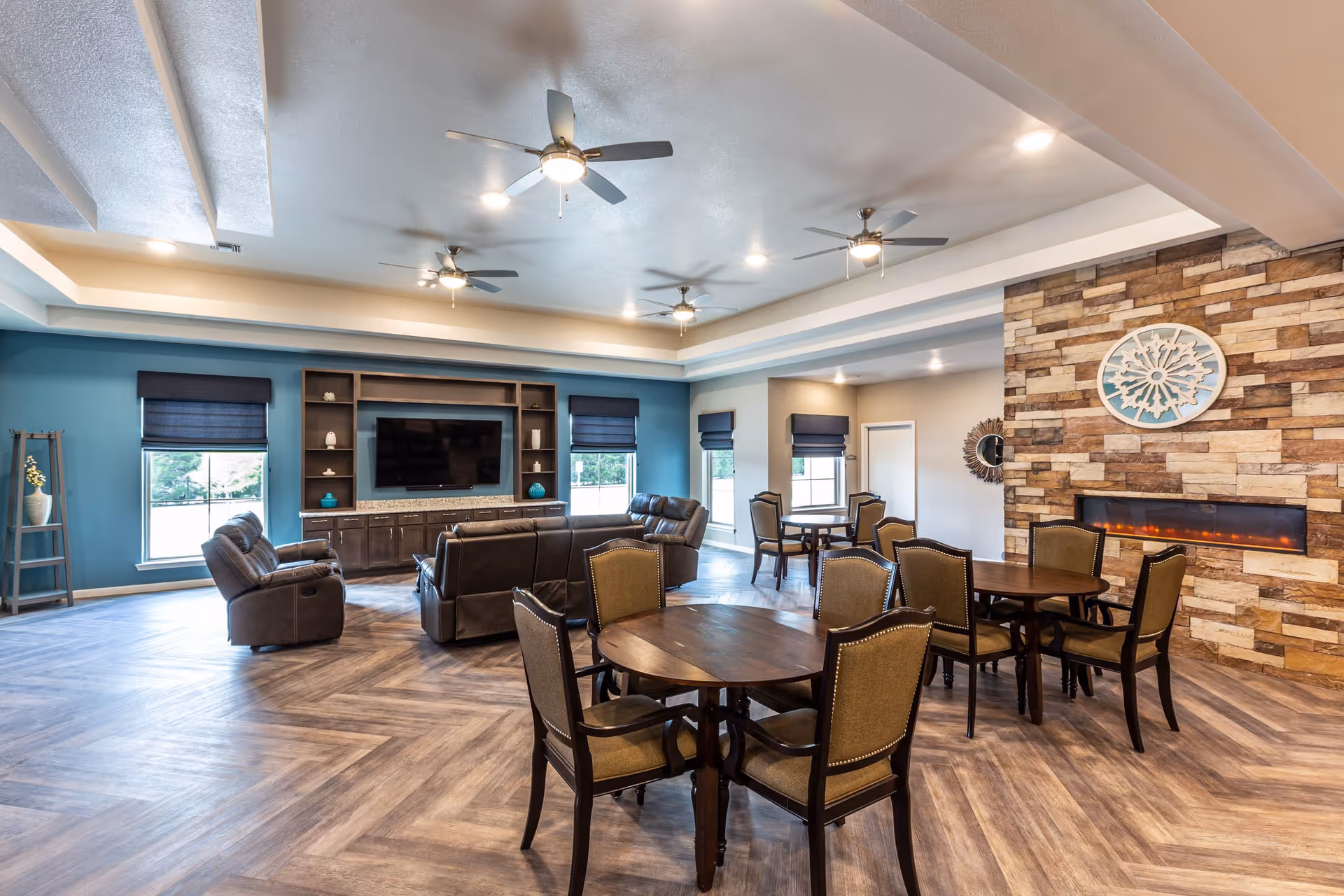 A spacious senior living common area with multiple round wooden tables and chairs, a seating area with leather recliners and sofas facing a large flat-screen TV mounted on a blue accent wall, and a modern electric fireplace set in a stone wall. The room has large windows with dark blinds, ceiling fans with lights, and wood-patterned flooring.