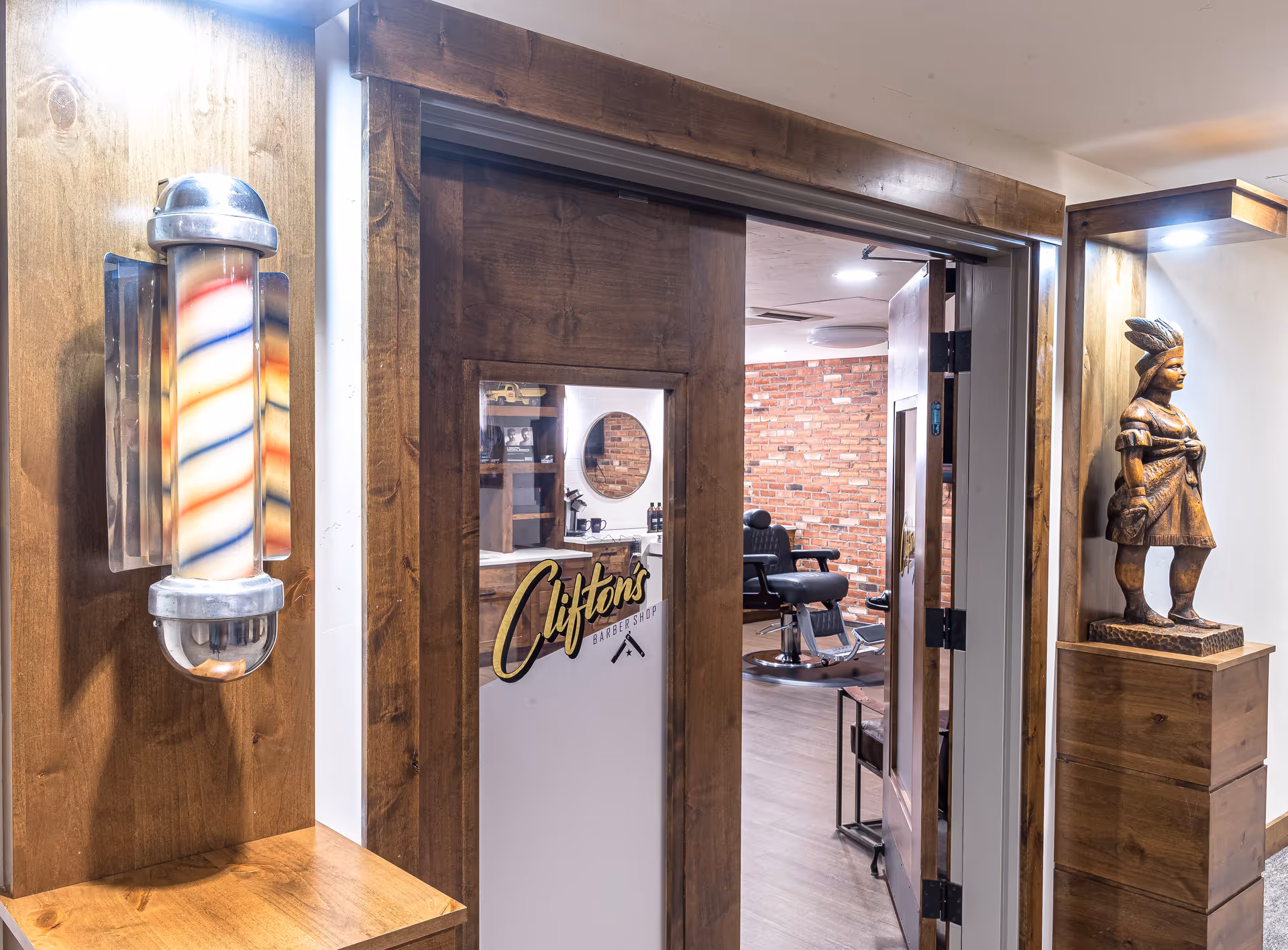 Interior view of a wooden-framed barbershop entrance with a lit barber pole, a 'Clifton's' decal on the door, and barber chairs visible inside.
