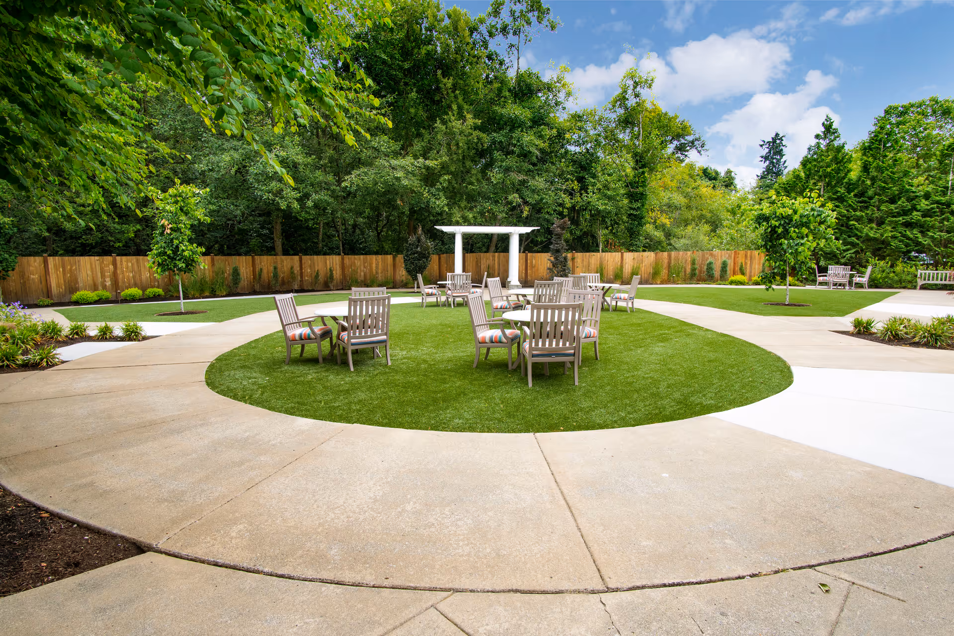 Outdoor seating area in a senior living facility with several wooden chairs and small tables arranged on a circular patch of artificial grass, surrounded by a concrete walkway. The area is bordered by a wooden fence and lush green trees under a partly cloudy blue sky.