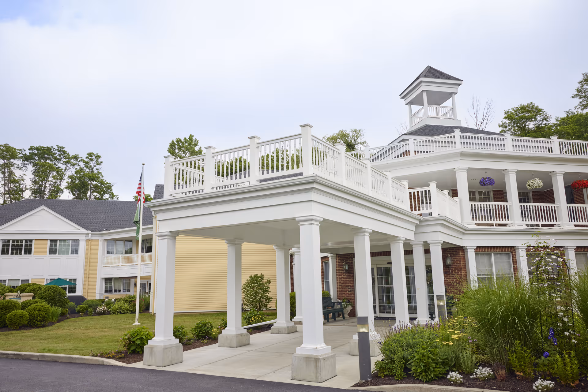 Exterior view of a senior living facility building with white columns and railings, a covered entrance, and a tower-like structure on the roof. The building is surrounded by greenery and landscaping, with an American flag and another flag visible near the entrance.