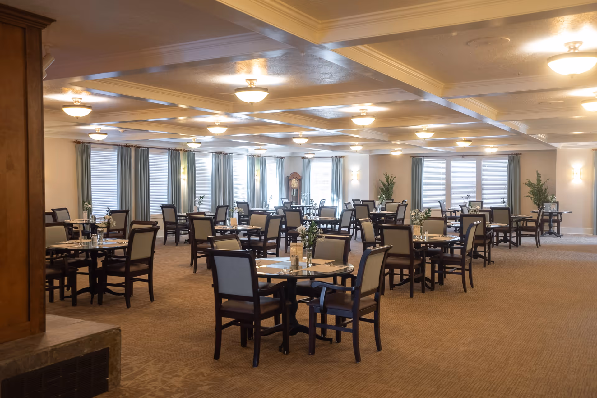 A spacious dining room with multiple round tables and chairs arranged neatly. Each table has a small floral centerpiece and place settings. The room has beige carpeting, light-colored walls, large windows with blue curtains, and ceiling lights evenly spaced across a coffered ceiling. There is a grandfather clock and some green plants in the background.