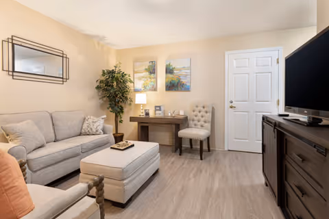A cozy living room with a beige sofa, matching ottoman, and an armchair with a peach cushion. There is a wooden desk with a lamp, chair, and decorative items against the wall. Two colorful paintings hang above the desk. A large flat-screen TV sits on a dark wooden cabinet opposite the sofa. The room has light wood flooring and neutral-colored walls.