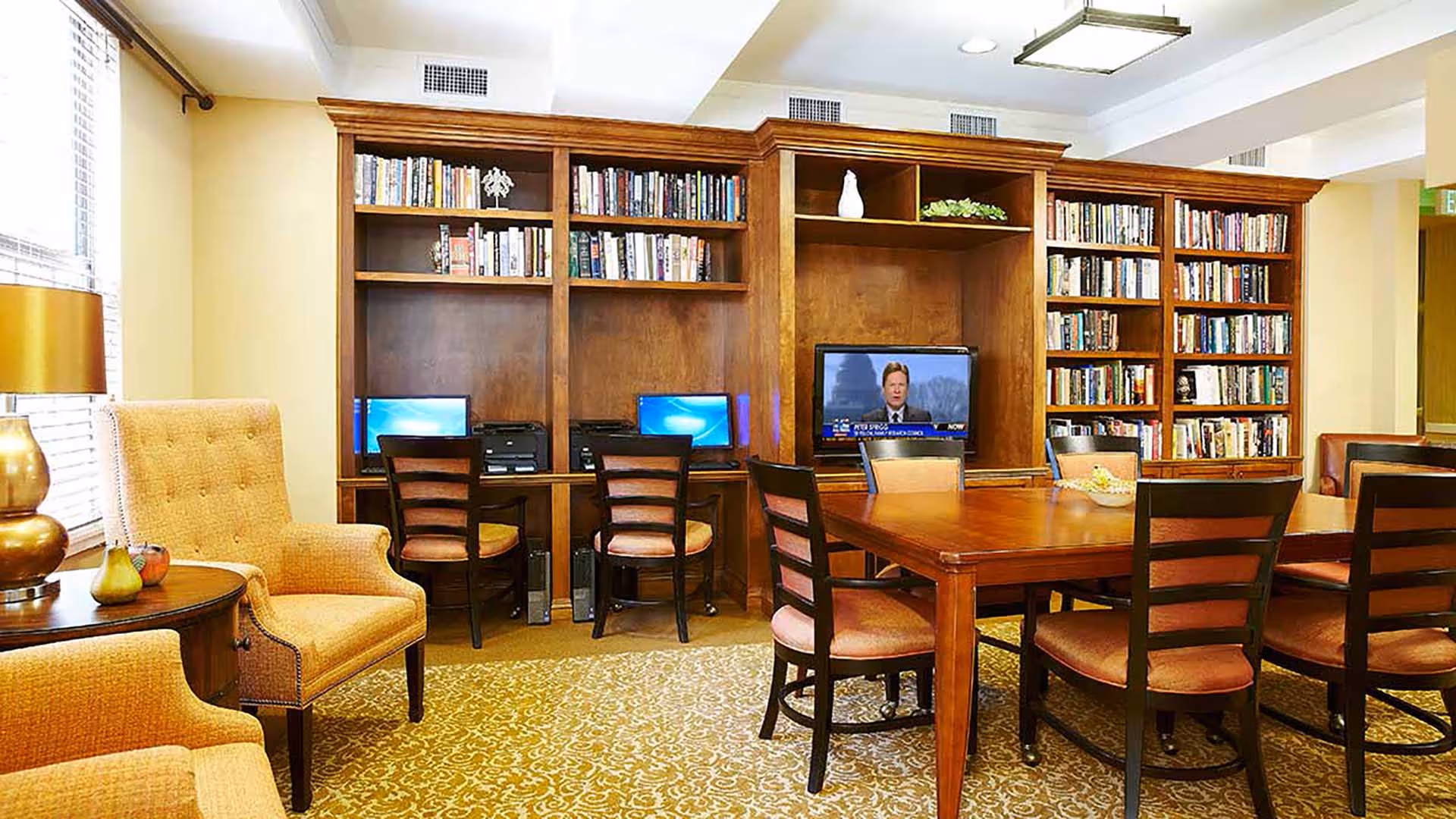A cozy common room featuring a large wooden table surrounded by chairs, a built-in wooden bookshelf filled with books, two computer workstations with monitors and printers, a television displaying a news program, and comfortable upholstered armchairs near a window with blinds.