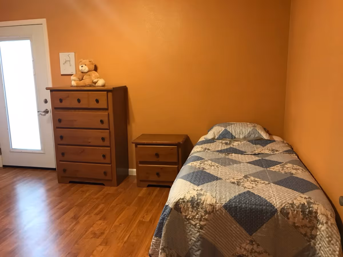 Small bedroom with a single bed against an orange wall, wooden nightstand and dresser topped with a teddy bear, and a frosted-glass door.