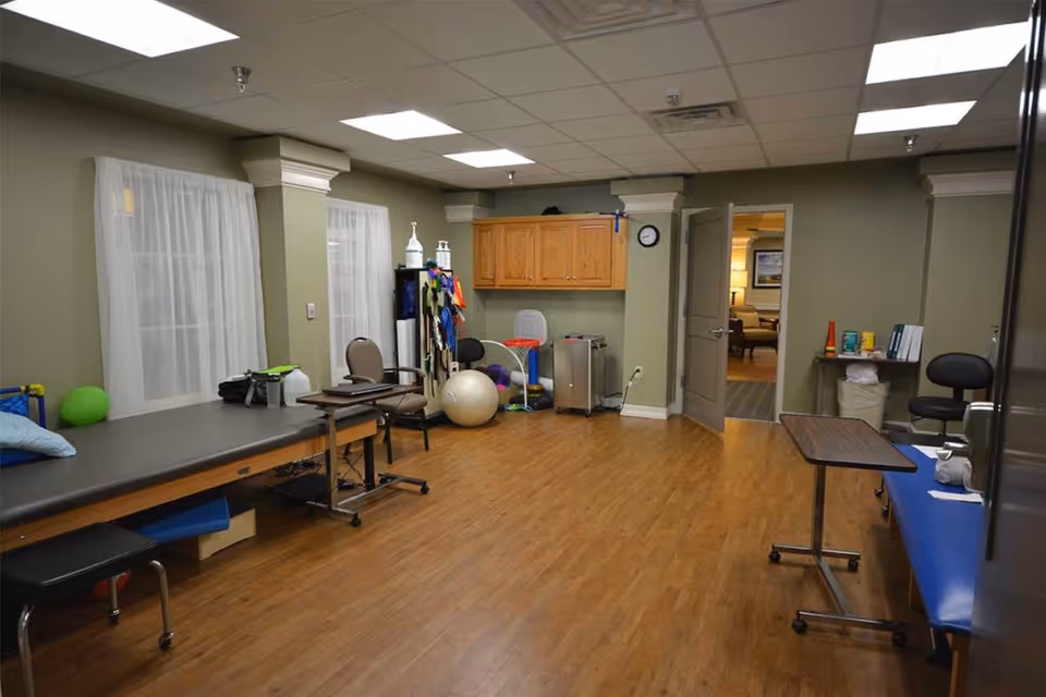 A spacious therapy or rehabilitation room with wooden flooring, two padded therapy tables, chairs, exercise balls, and various therapy equipment. The room has green walls, white sheer curtains on the windows, and a door leading to another room with seating visible.