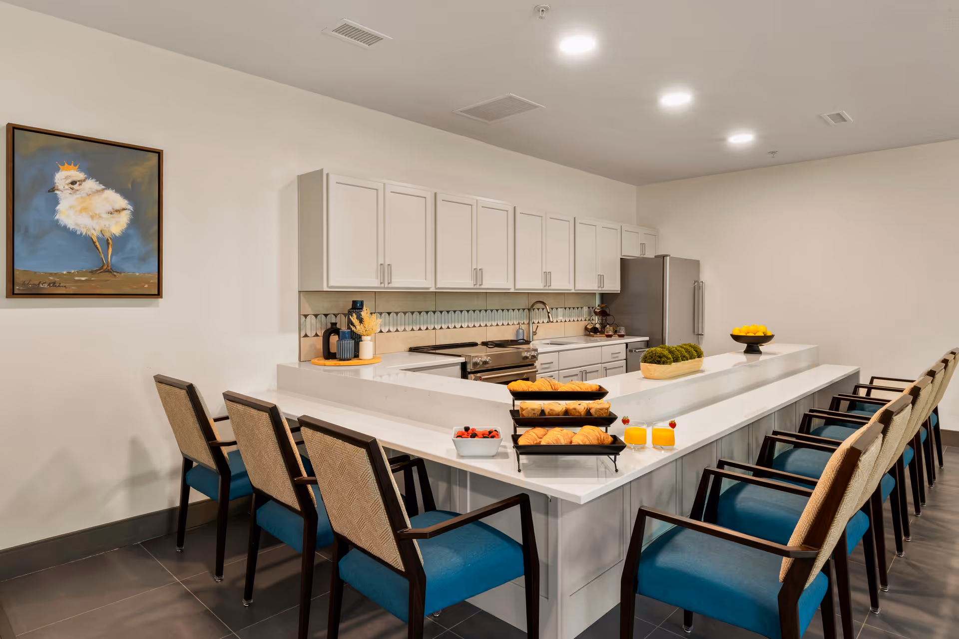 Bright communal kitchen with a long white island, blue-upholstered chairs, and trays of pastries and juice.
