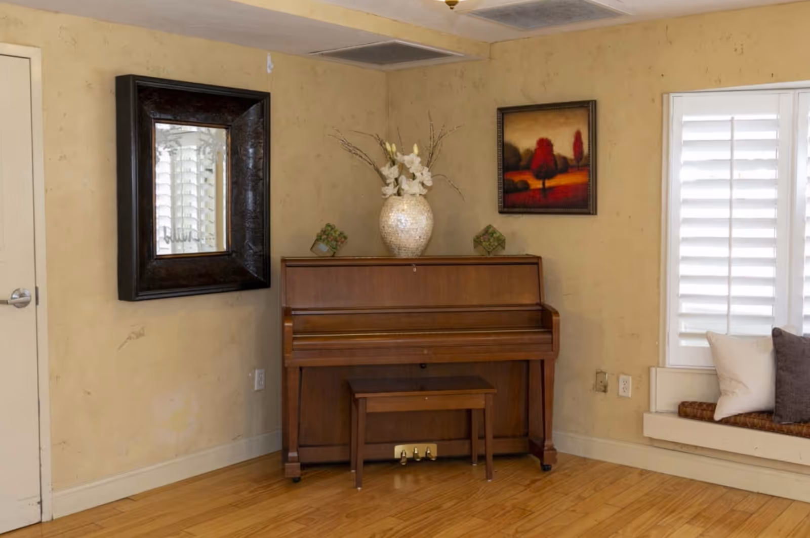 A cozy corner of a room featuring a wooden upright piano with a matching bench. On top of the piano is a decorative vase with white flowers and two small green plants. To the left of the piano is a large dark-framed mirror hanging on a beige wall. To the right, there is a window with white plantation shutters and a cushioned window seat with pillows. A framed painting of trees with red and orange hues hangs above the piano.