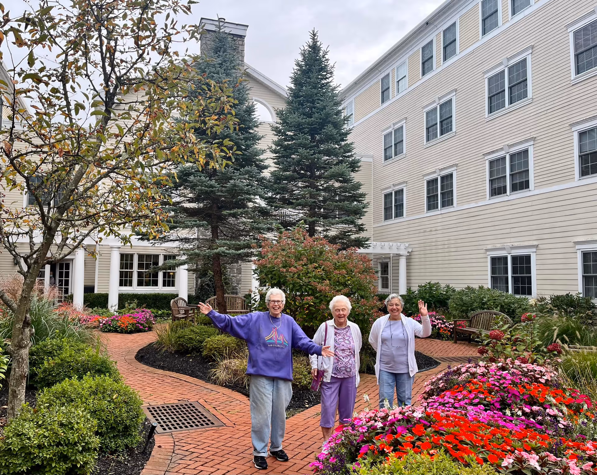 Three elderly women standing and smiling in a landscaped courtyard with colorful flowers, bushes, and trees, surrounded by a multi-story beige building with white trim and multiple windows.