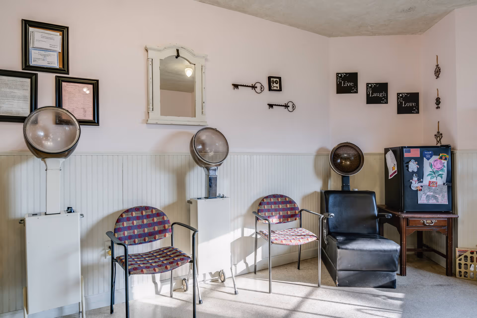 A small salon area with three vintage hair dryers, two patterned chairs, and one black salon chair. The walls are decorated with framed certificates, a white-framed mirror, decorative keys, and three small wall plaques with the words 'Live', 'Laugh', and 'Love'. There is a small table with a mini fridge covered in magnets and drawings.