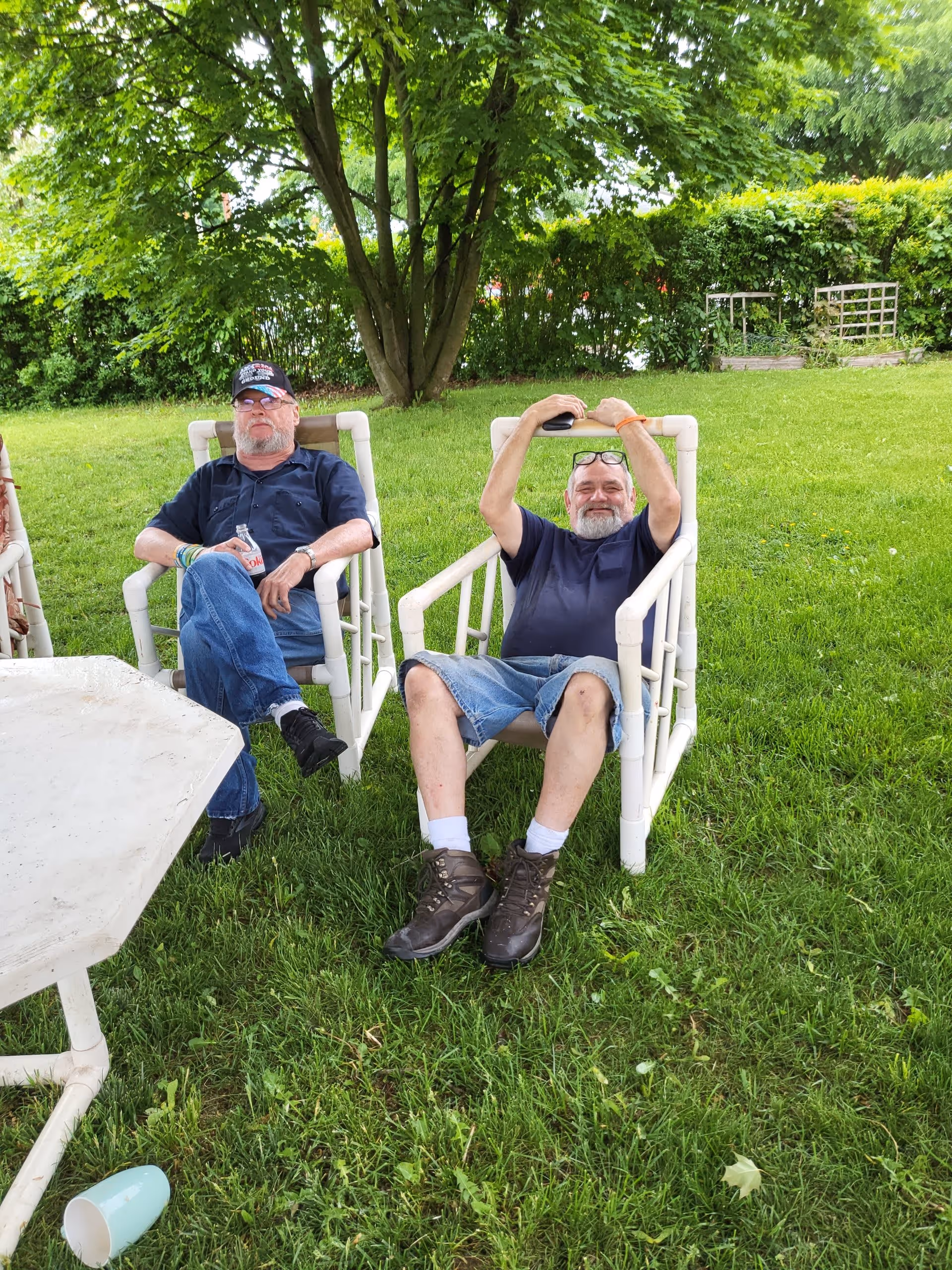 Two older men sitting outdoors on white plastic chairs on green grass under a tree. One man is wearing a black cap, glasses, a dark shirt, and jeans, holding a bottle. The other man is wearing a dark t-shirt, denim shorts, and hiking boots, with glasses resting on his forehead and his arms raised behind his head. There is a white table and a blue cup on the grass nearby, with bushes and trees in the background.