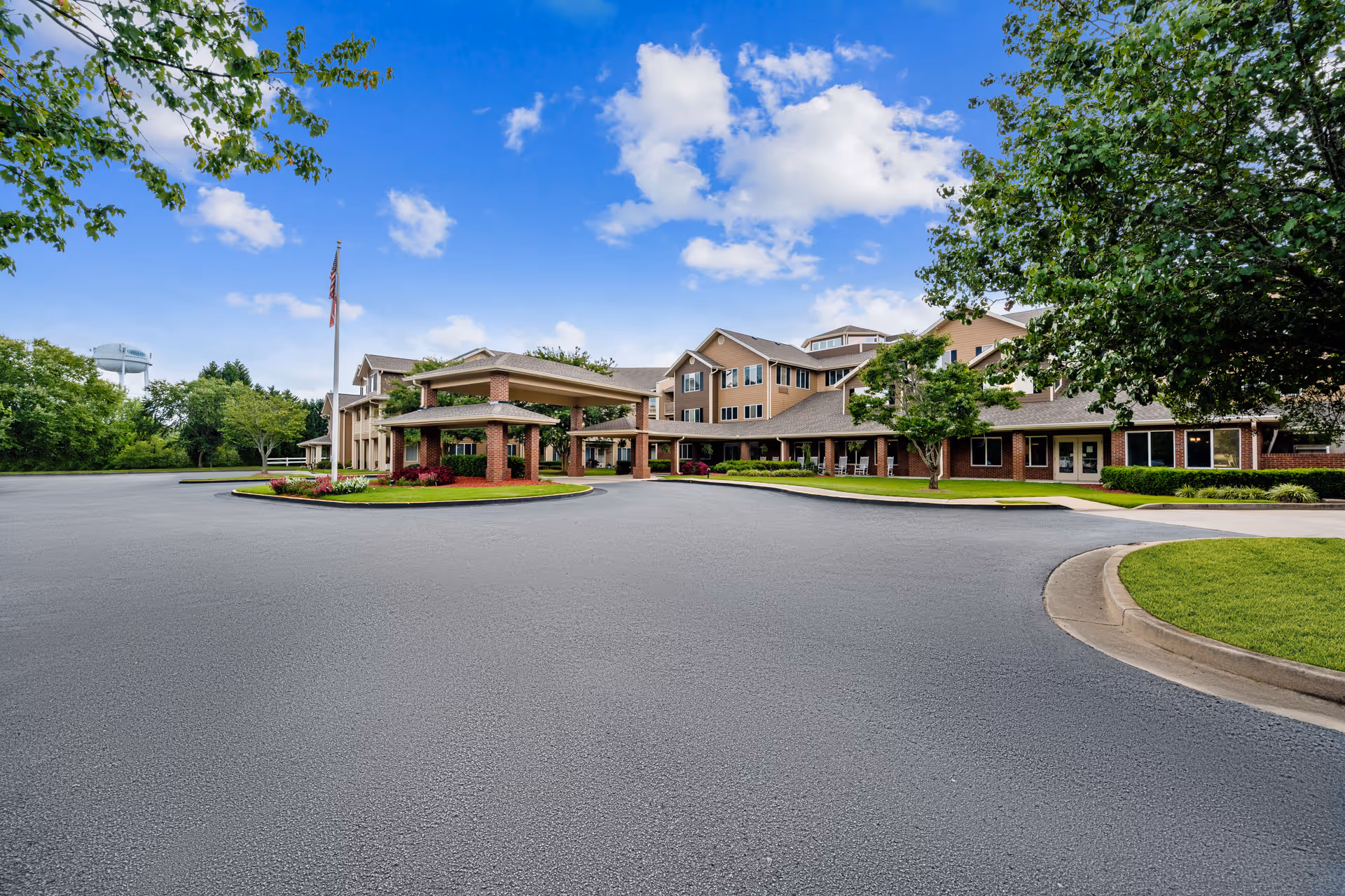 Exterior view of Holiday Pinegate senior living facility showing a large driveway with a covered entrance, surrounded by well-maintained landscaping, trees, and a clear blue sky with some clouds.
