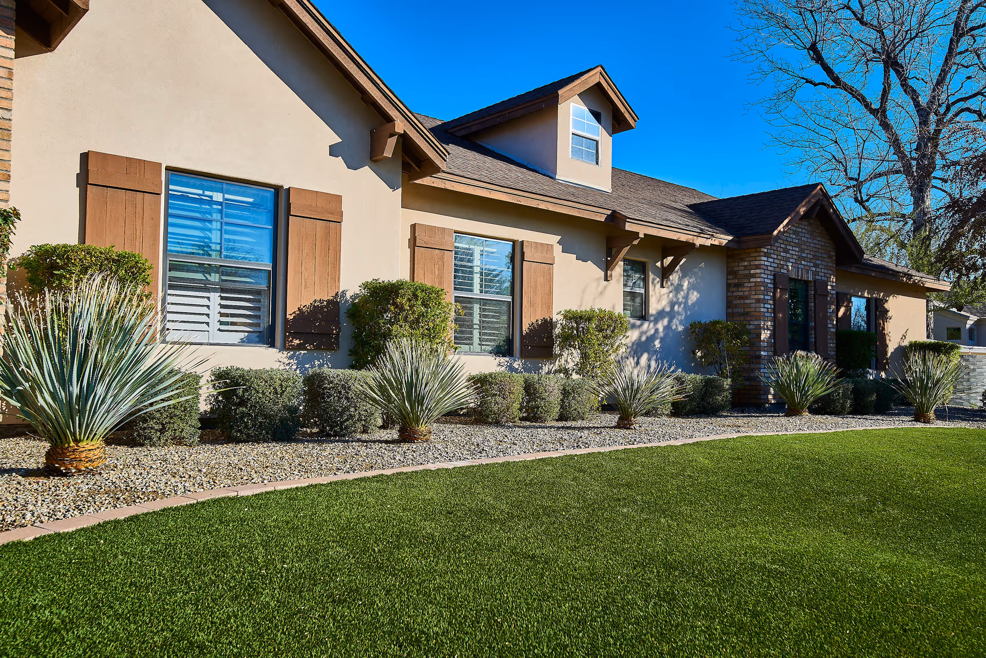 Exterior view of a single-story building with beige walls, brown wooden shutters on the windows, and a well-maintained front yard featuring green grass, small bushes, and desert plants under a clear blue sky.