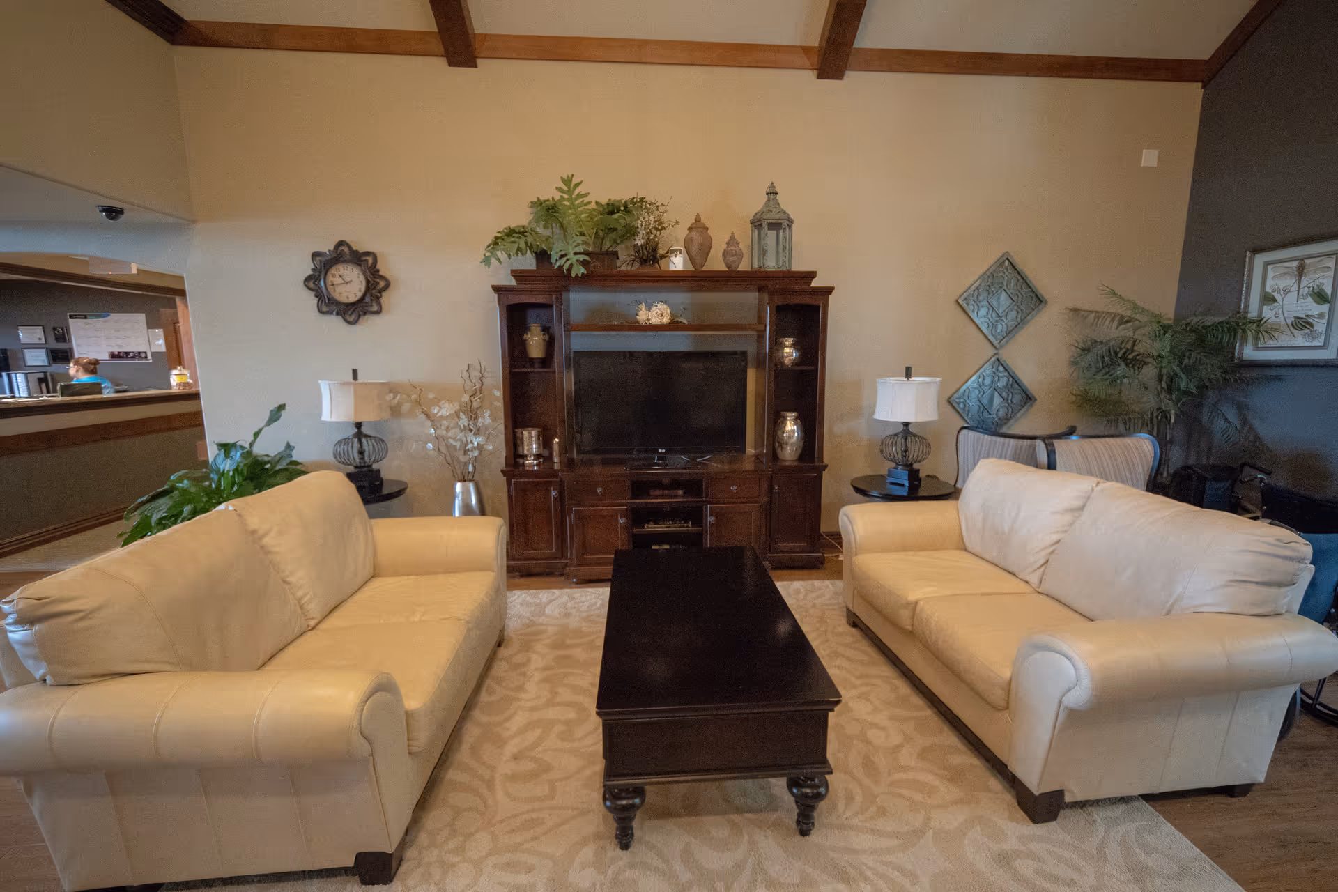 A cozy living room with two beige leather sofas facing each other across a dark wooden coffee table. Behind the coffee table is a dark wooden entertainment center with a flat-screen TV, decorative vases, and plants on top. The room has a beige patterned rug, two side tables with lamps, wall decorations including a clock and framed artwork, and a potted plant in the corner.