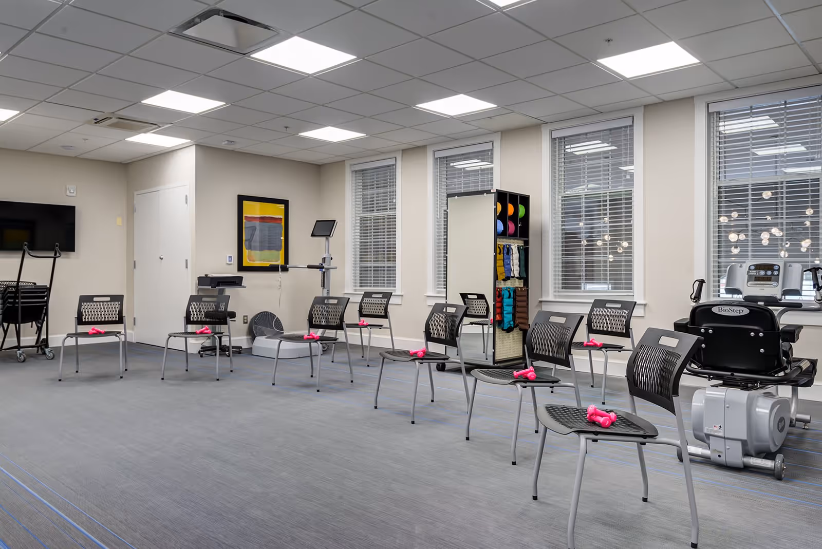 A spacious exercise room with multiple chairs arranged in a semi-circle, each chair holding a pair of pink dumbbells. The room has large windows with blinds, exercise equipment including a stationary bike and a step machine, and a rack with colorful exercise balls and bands. The walls are light-colored, and the ceiling has recessed lighting panels.