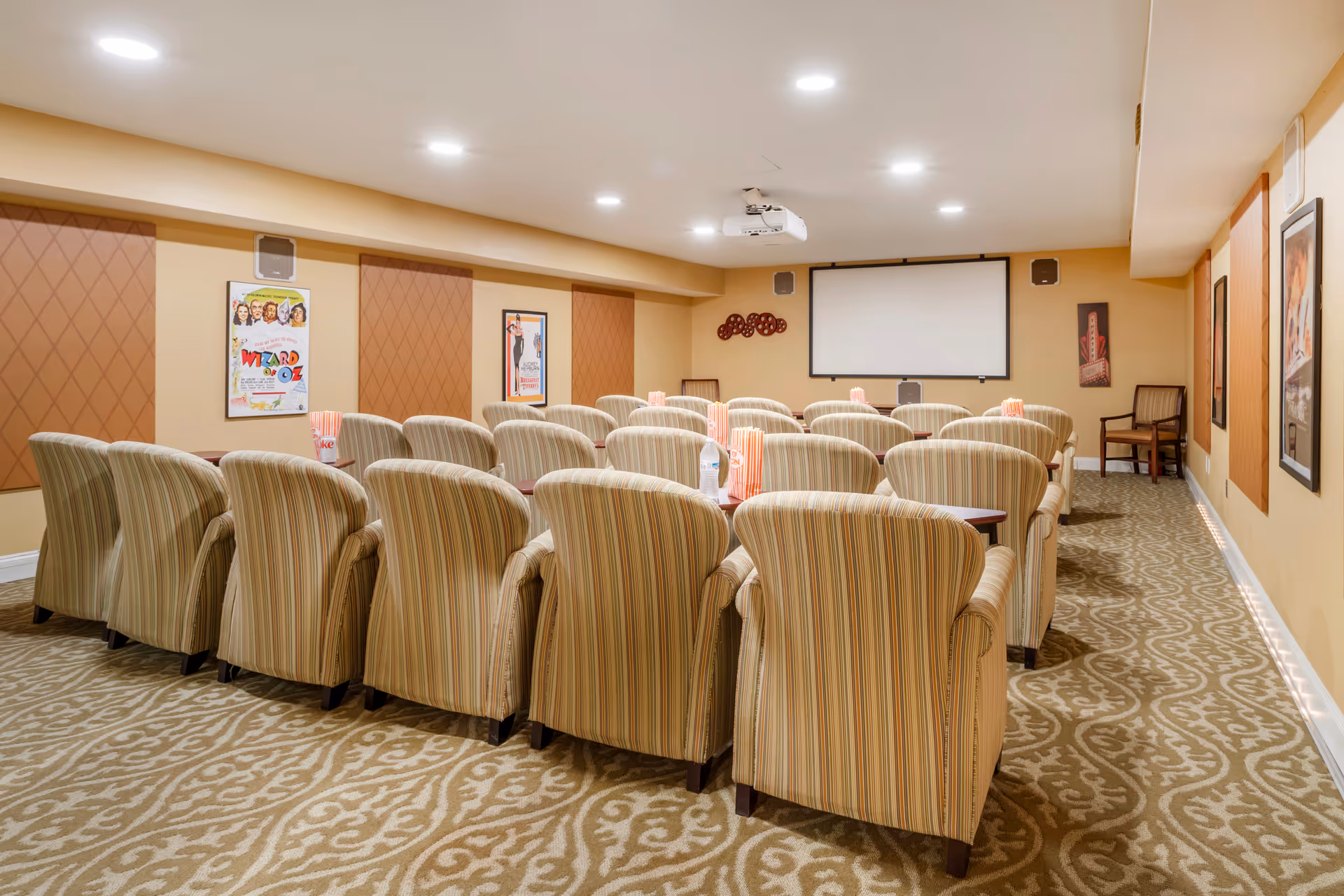 A cozy movie theater room with multiple rows of striped upholstered chairs facing a large projector screen. The room has beige walls adorned with classic movie posters and a patterned carpet. Popcorn containers and a water bottle are placed on small tables between the chairs.