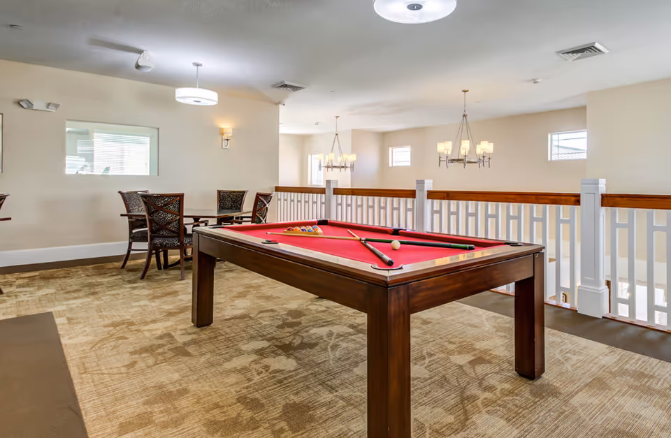 A bright indoor recreational area featuring a pool table with a red felt surface and pool cues resting on it. In the background, there are several chairs and a small table near a window. The room has beige walls, carpeted flooring, and modern light fixtures hanging from the ceiling. A white railing separates this area from a lower level.