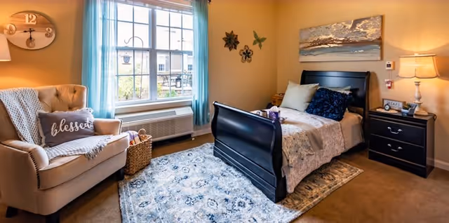 Cozy bedroom with a dark wood sleigh bed, patterned rug, armchair by a window with blue curtains, and a nightstand with a lamp.
