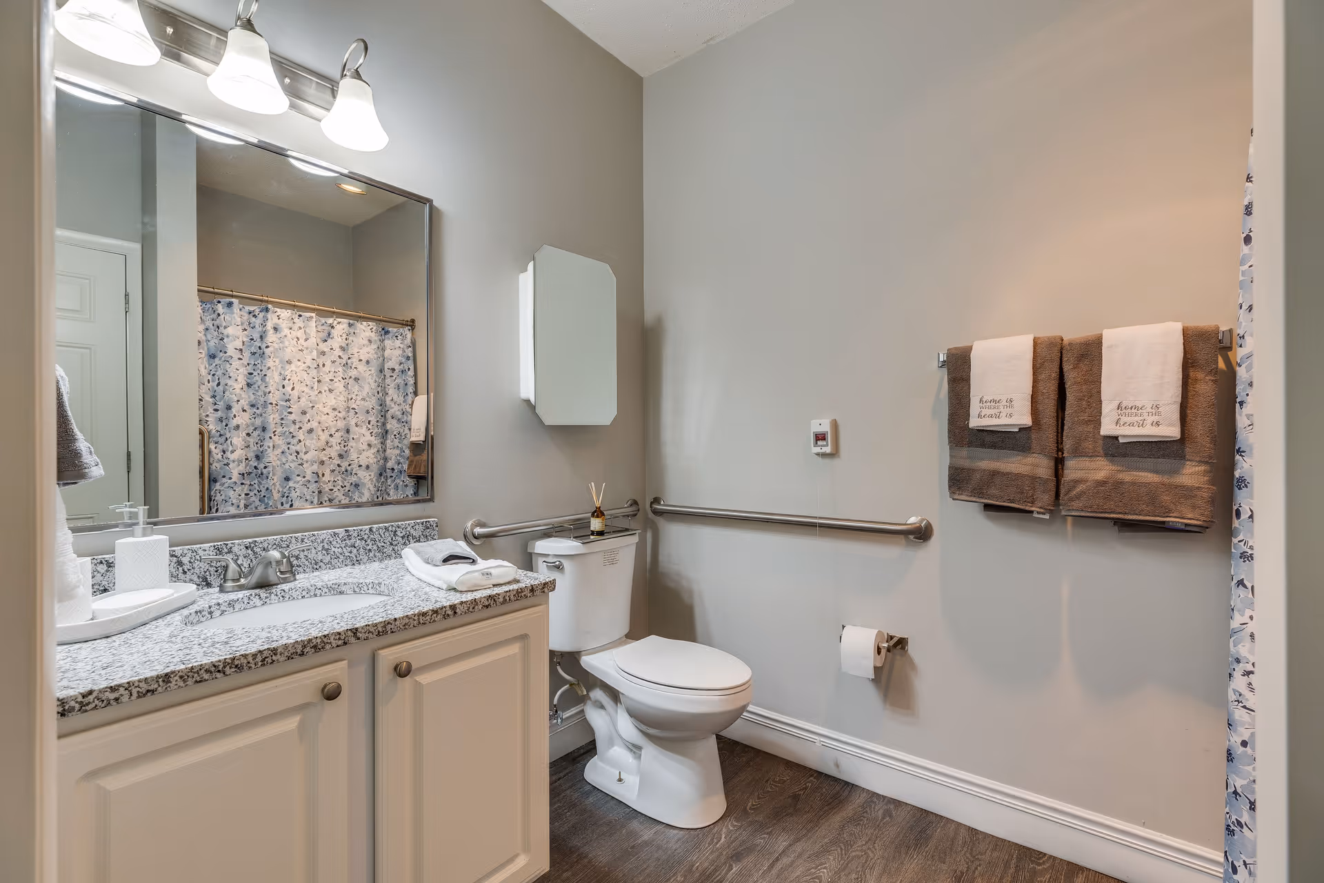 A clean bathroom with a white toilet, a granite countertop with a sink, a large mirror above the sink, and a shower with a floral-patterned curtain. There are two brown towels with white hand towels hanging on a towel rack on the wall, and a grab bar installed next to the toilet. The floor is wood-style, and the walls are painted light gray.