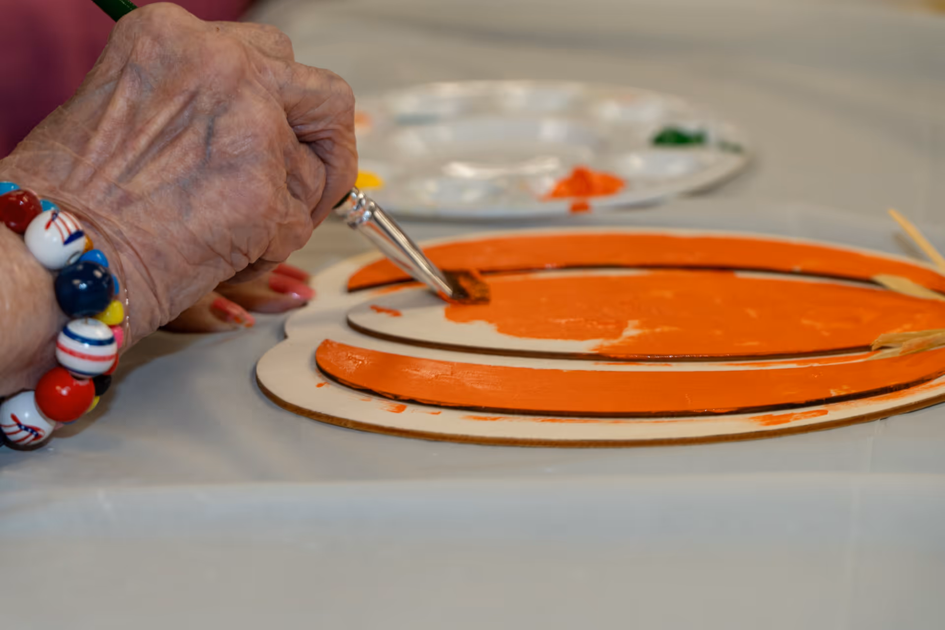 Close-up of an elderly person's hand painting an orange pumpkin-shaped wooden craft with a paintbrush on a table covered with a protective sheet. A paint palette with various colors is visible in the background.