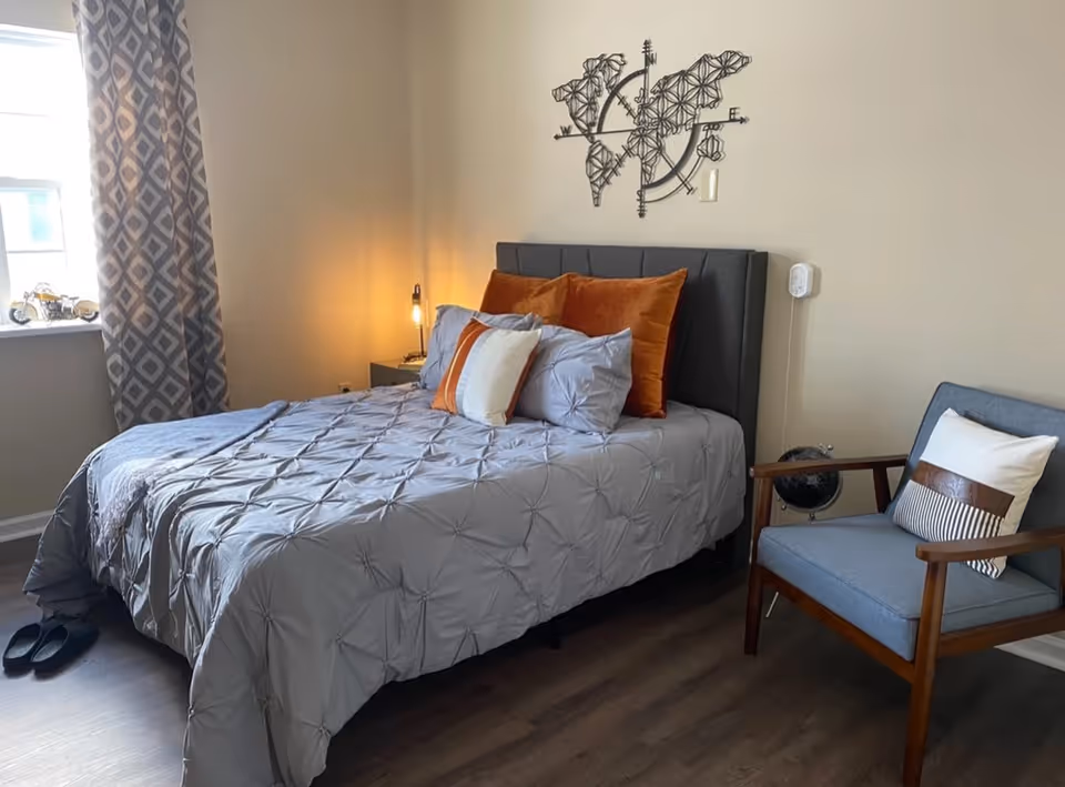 A neatly made bed with gray bedding and orange and white pillows in a bedroom. There is a patterned curtain on the window to the left, a small nightstand with a lamp beside the bed, a decorative metal wall art above the bed, and a blue armchair with a pillow to the right. The floor is wooden.