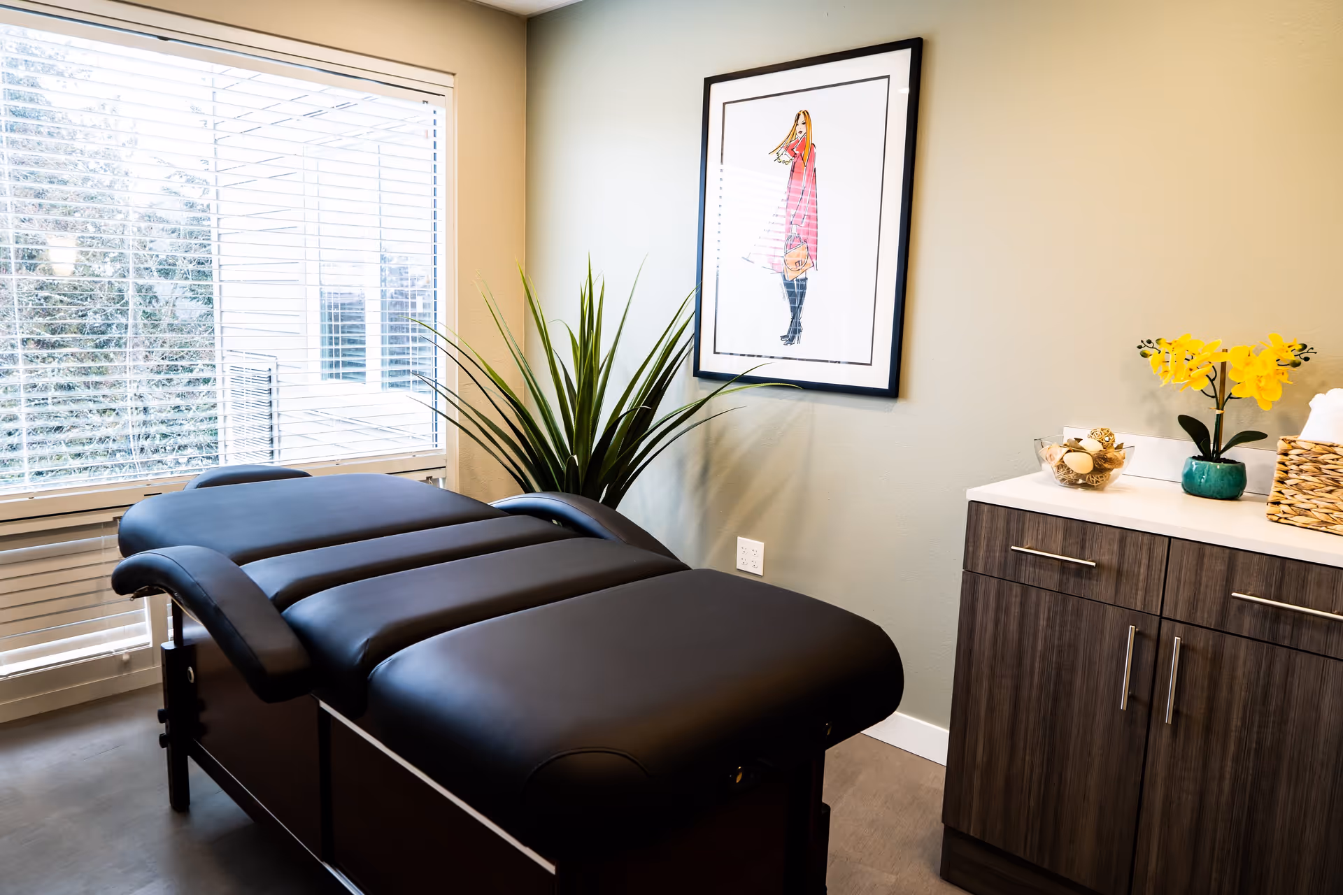 A modern treatment room with a black padded massage or therapy table, a large window with white blinds, a tall green plant in the corner, a framed fashion illustration on the wall, and a wooden cabinet with a white countertop holding a yellow orchid plant and decorative items.
