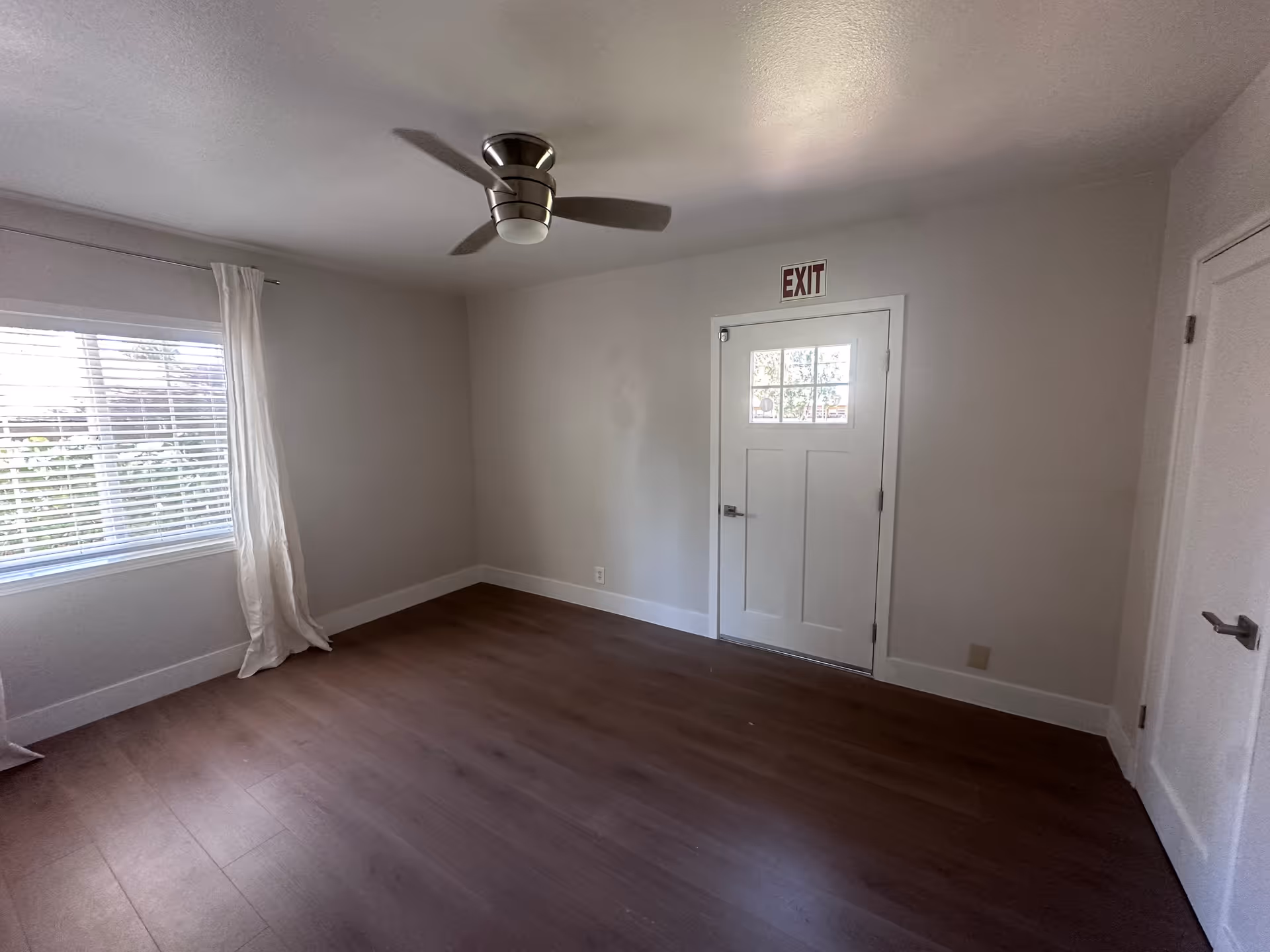 A bright empty room with a window, ceiling fan, and an exit door.