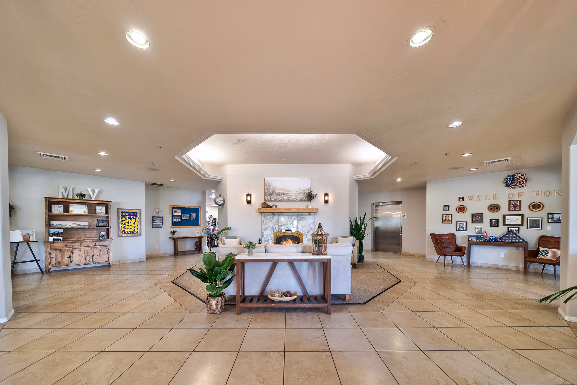 Spacious senior living facility common area with tiled floors, a central seating arrangement around a fireplace, plants, and wall decorations including a 'Wall of Honor' with framed photos and memorabilia.