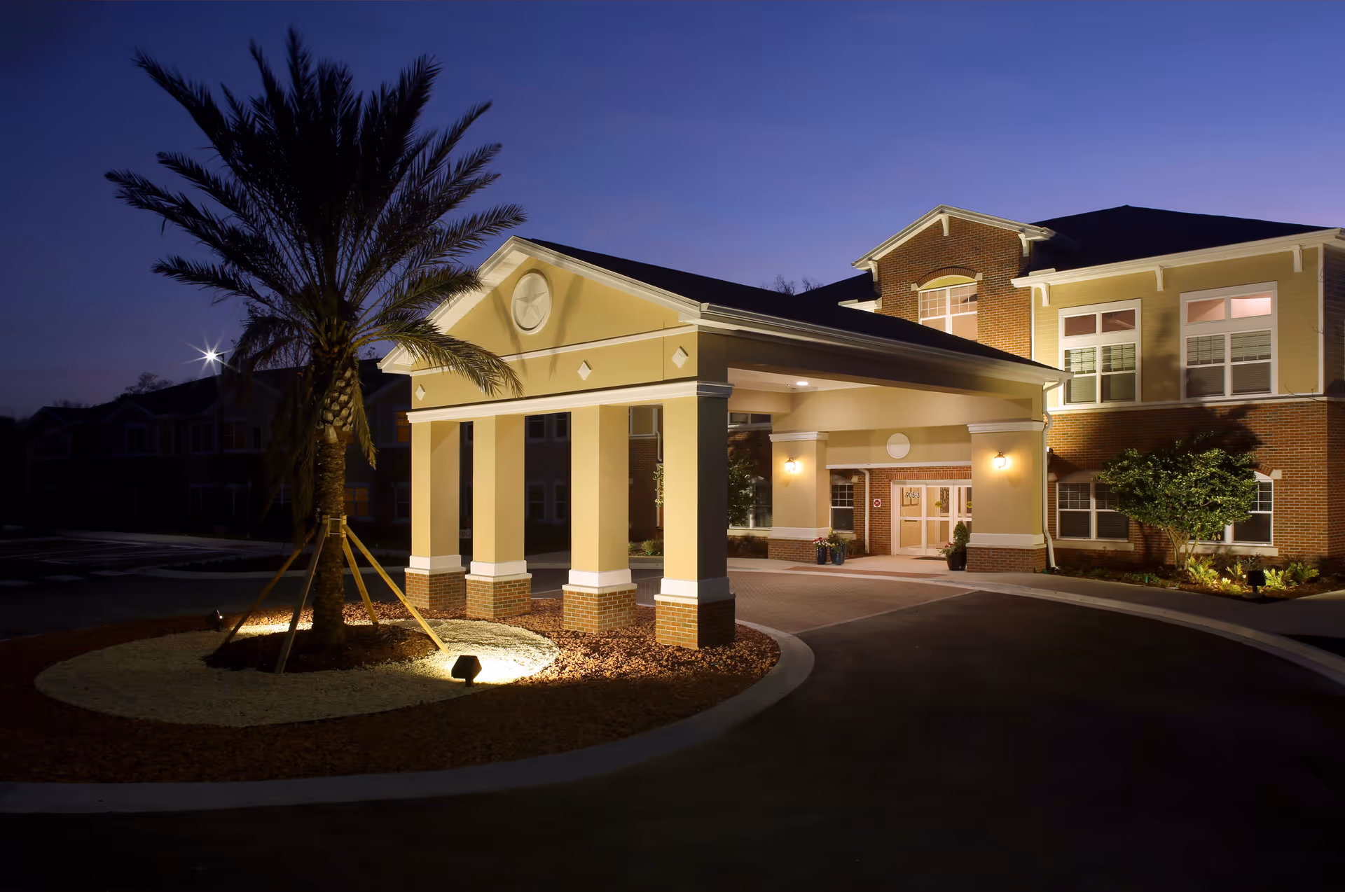 Exterior view of Anthem Lakes senior living facility at dusk, showing the well-lit entrance with a covered driveway, a palm tree in a landscaped circular bed, and the building's brick and beige facade with multiple windows.
